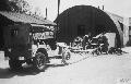 A Jeep which has been jacked-up to enable one of the front wheels to turn freely, in order to power a bench saw.Two members of groud crew of the 55th Fighter Group are using the saw to cut trees.